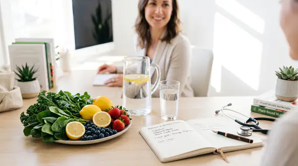 A glass of water, fresh fruits, and leafy greens arranged on a light wooden table alongside a notebook, pen, and stethoscope, representing safe dietary changes and nutrition consultation.