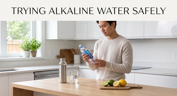 A person in a modern kitchen holds and reads the label of an alkaline water bottle, with lemons, limes, and a glass of water on the counter, next to text overlay "TRYING ALKALINE WATER SAFELY".